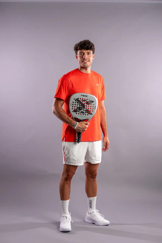 Full-length studio photograph of a player in orange NOX clothing holding the AT10 Genius Attack 12K Alum XTREM 2027 racket