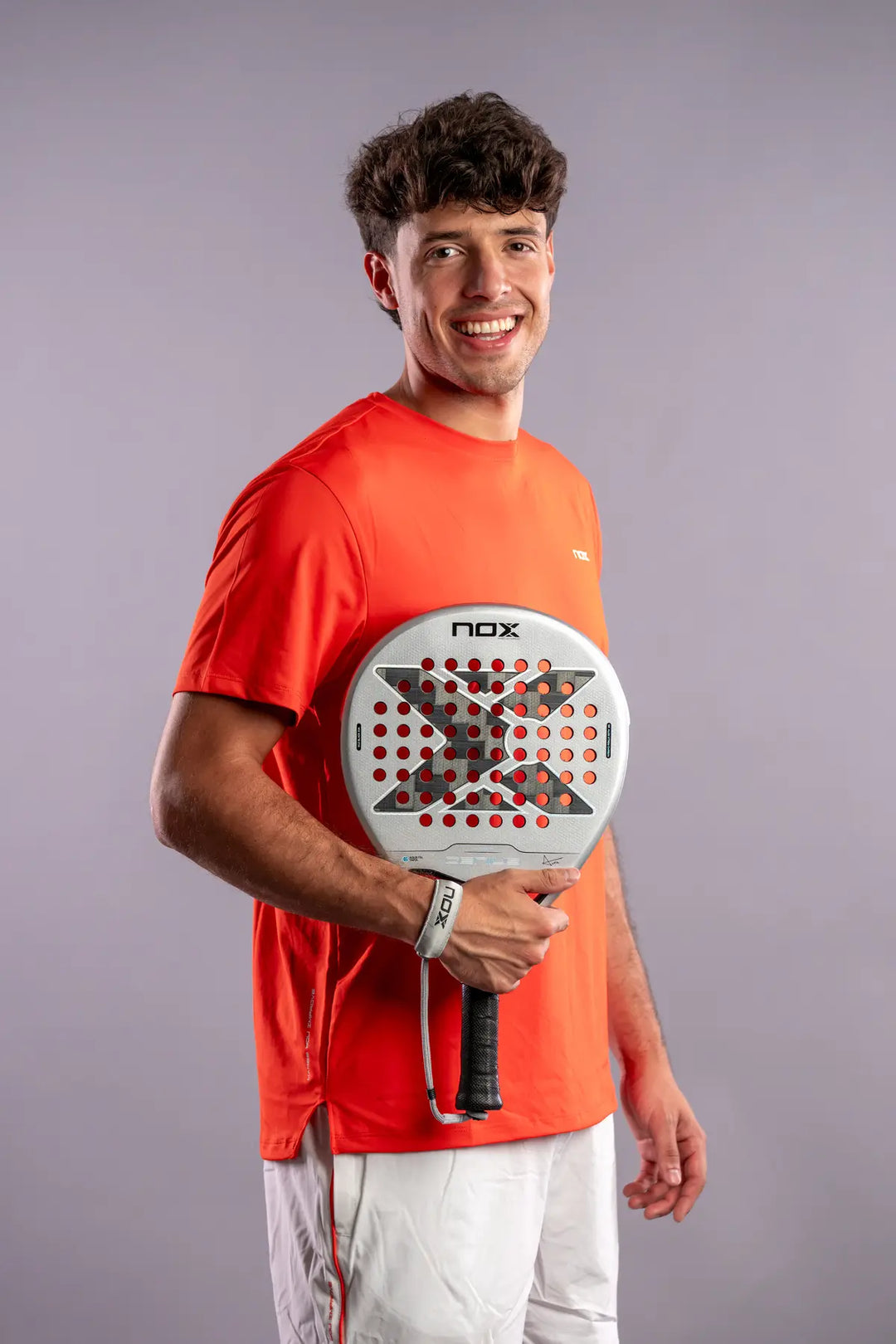 Agustín Tapia holding the silver NOX Genius 12K Alum XTREM 2027 padel racket in front of his orange NOX shirt