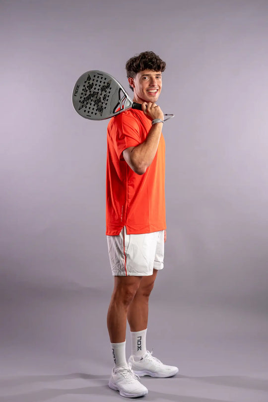 Agustín Tapia holding the NOX Genius 12K Alum XTREM 2027 padel racket over his shoulder in an orange NOX shirt