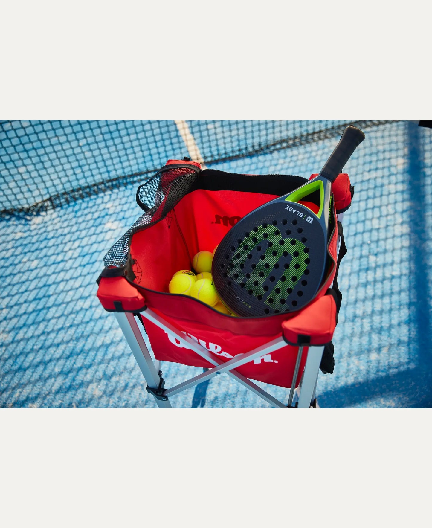 Wilson Padel X3 balls in a red training basket with padel racket on an outdoor padel court
